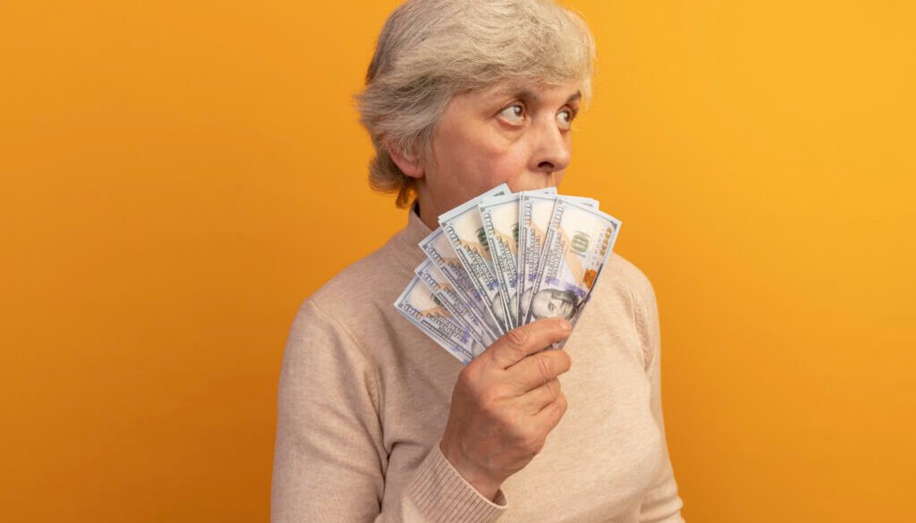 old woman wearing creamy turtleneck sweater standing in profile view holding money looking up from behind it isolated on orange background with copy space