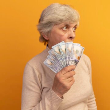 old woman wearing creamy turtleneck sweater standing in profile view holding money looking up from behind it isolated on orange background with copy space