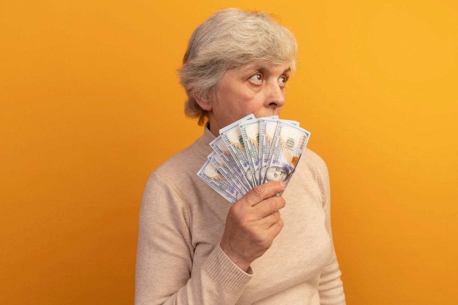 old woman wearing creamy turtleneck sweater standing in profile view holding money looking up from behind it isolated on orange background with copy space